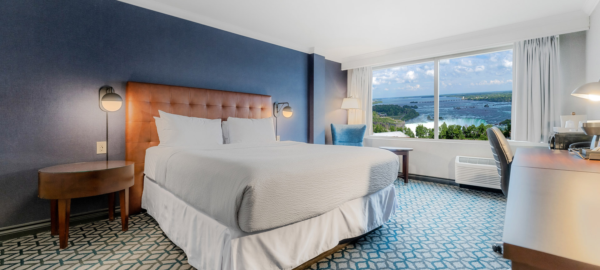 A hotel room with a king bed with white linen, a large window with a view of Niagara Falls and a spacious desk for work or study purposes.